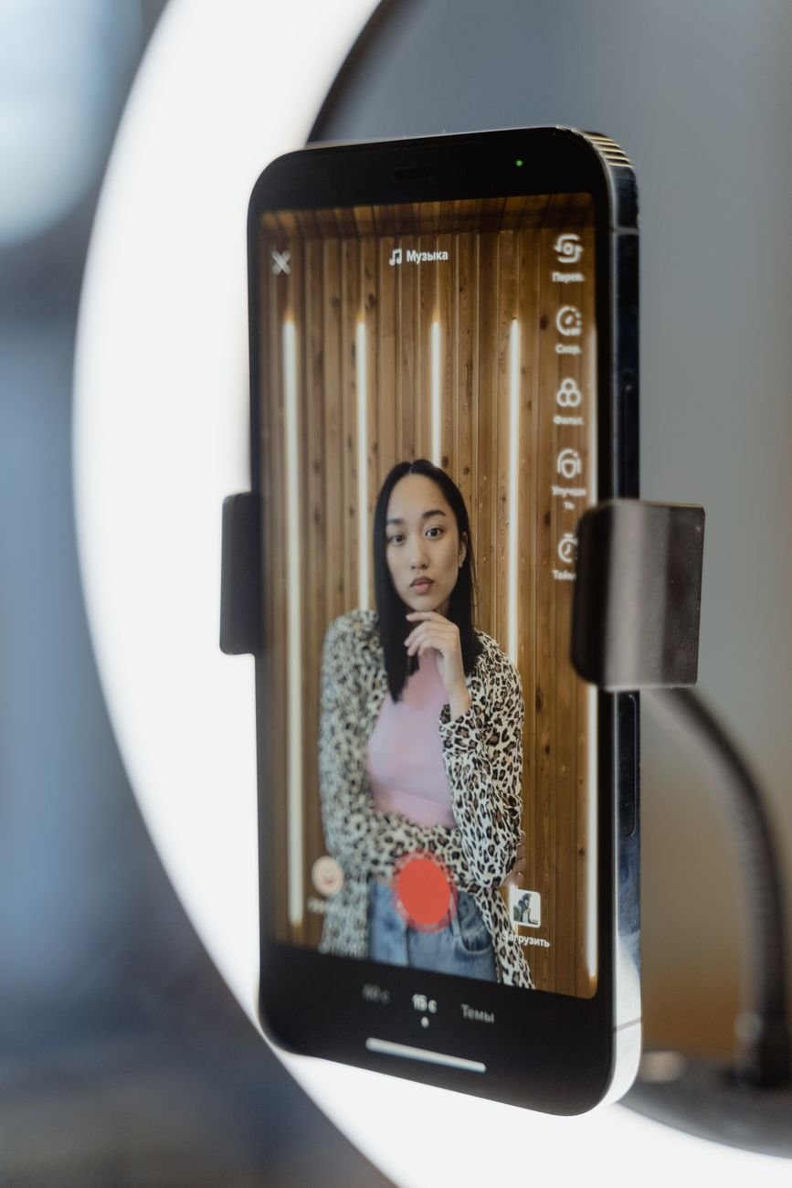 Woman recording a TikTok video using a smartphone and ring light indoors.